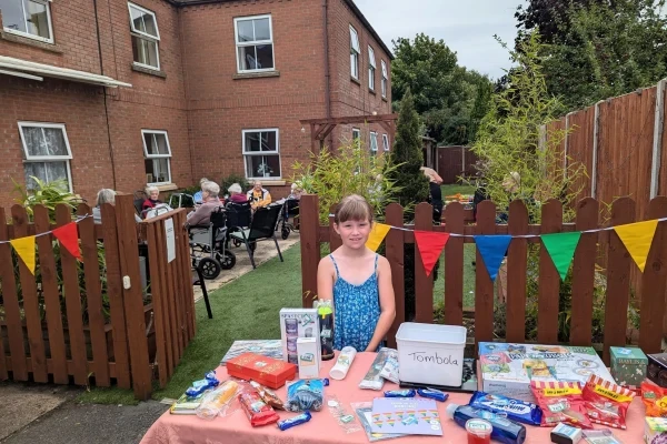 A young visitor running a stall at a garden fête, with residents in the background